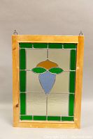 Front view of rectangular stained glass window panel with wooden frame and brass hanging hardware. Shows geometric floral design in green, blue, and amber glass with clear textured glass background.