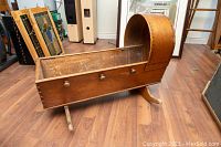 Side view showing the length and curved canopy end on wooden cradle with three knobs on side and rocker legs, positioned on wood floor indoors among other vintage items.