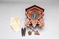 Front view of the hand-carved wooden cuckoo clock, showing detailed leaves and bird with clock face and hands, pine cone weights, chain, pendulum, and instruction sheet.