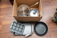 Frying pans, covered pot, rectangular pans, and mini muffin tins arranged inside a box, shown on wooden floor.
