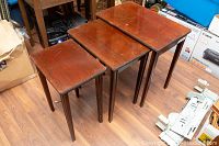 Three wooden nesting tables with rectangular tops and straight legs arranged next to each other on wooden floor with some surrounding clutter. Tables show medium wear.