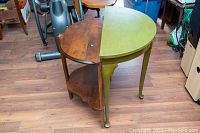 Two vintage demi-lune tables side by side, one with green paint and brass feet, the other natural wood with wear.