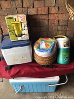 Overview of camping supplies showing coolers, stacked paper plates, beverage cooler, and Empire electric hot cup in original box.