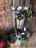 Golf bag holding multiple right-handed Spectra golf clubs, box of golf balls, and Bennington pull cart seen against brick wall on concrete floor.