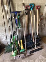 Photo showing three shovels, two brooms, three rakes including a metal leaf rake and a bright green plastic rake, and a yellow/green weed trimmer.