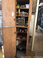 Full view of tall wooden cabinet with multiple shelves showing assorted hardware including small boxes, jars containing nails, screws, and other small items.