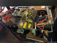 Photo showing various tools including a caulking gun, glue gun, measuring tapes, drill bits, and power cords on a shelf.