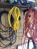 Three extension cords hanging coiled on a wall mount in a garage environment: black, yellow, and red colors visible.