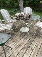 Overview of round glass table on wood deck with four striped cushioned metal chairs and folded umbrella on top.