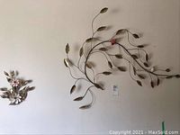 Wide view showing the large metal leaf and bird wall art with the small shell flower art to the left on a cream wall