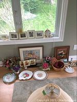 Full view of lot items on floor by window showing floral arrangements, chinaware, baskets, paintings, plaques, and souvenirs.