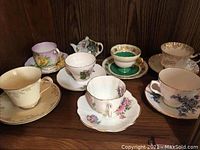 Full view of the seven china teacups and saucers on wooden surface, showing varied patterns and colors.