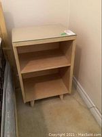 Wood end table with protective glass top, two shelves, light wood finish, positioned next to a mattress.