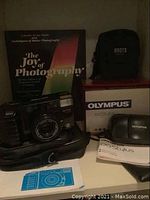 Minolta AF-Tele camera front with lens and autofocus visible alongside Roots camera bag, Olympus box, manual, and photo albums behind.