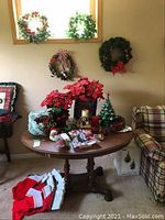 Wide view showing four Christmas wreaths mounted on the wall above a wooden table. On the table are several faux poinsettia plants, Christmas candles, and stocking items on the floor nearby.