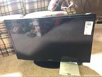 Front view of the Samsung 32-inch LED TV on stand with remote and manual visible on carpet.