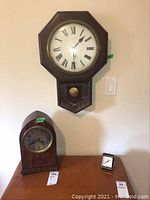 Photo showing the three clocks: a large wall hanging clock with Roman numerals mounted on the wall, a wooden mantel clock with Arabic numerals, and a small travel clock placed on a table.