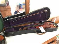 Full view of violin in open black hard case with bow inside, purple velvet lining.