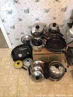 Photo shows collection of various pots, pans, lids, and a large roaster pan stacked on the floor, displaying the different sizes and types of cookware.