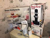 Photo showing boxed appliances: Braun Multipractic hand blender, Starfrit electric peeler, Black & Decker 2-speed super chopper, and Braun juicer boxes stacked on a corner table with some kitchen appliances visible.