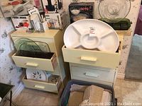 Two pale yellow painted wood cabinets with four drawers each, drawers partially open showing glass trays and dishes inside. Cabinets show wear and chipping paint.