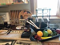 View of kitchen countertop showing wooden cheese tray with ceramic plate, wine glasses with colored jewels, wine rack, faux fruit in black bowl, wooden salad bowl, and slate coasters.