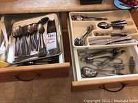 First image showing two open drawers with trays containing various spoons, forks, knives, tongs, and specialty serving utensils in stainless steel with decorative handles.
