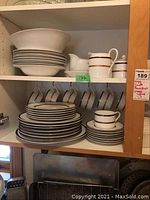 Cabinet shelf with stacked white plates with gold/brown bands, bowls, cups hanging on hooks, cream and sugar dishes