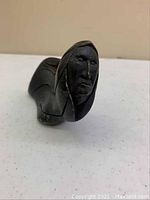 Front and side view of a small black wooden carving depicting a stylized human figure with headdress.