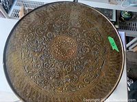 Close-up photo of the ornate hand hammered brass tabletop showing floral and wave patterns with colored inlay details.