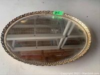 Photo of an oval glass dresser tray with a decorative metal rim, size approximately 16x12 inches, showing the reflective glass surface and ornate edge.