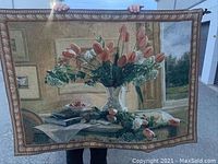 Tapestry held by a person showing full front view with floral still life design featuring tulips and roses in a vase on a table against a detailed interior background