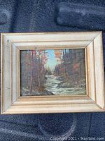 Framed oil on board painting depicting a wooded landscape with a flowing stream, showing overall condition and framing.