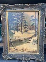 Front-facing image of a framed oil painting showing a winter landscape with snow-covered ground, trees, and a small wooden bridge. Frame is ornate with floral and scroll details, shows some wear including a chip.