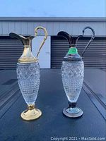 Pair of Greek style wine carafes with metal tops, handles and bases, one gold-toned and one silver-toned, shown side by side