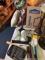 Wide view of desk lamp, JBL speakers, paper punch, clock radio, alarm clock, rubber bands, and bulbs packed on table.