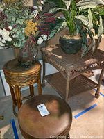 Photo showing square wicker table with faux plant in black pot, smaller round wicker table, and faux floral arrangement in clear glass container.