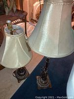 Two metal table lamps of different heights with cream fabric shades, visible wear and scratches.