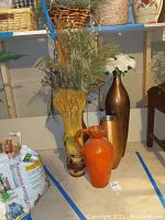 Five vases and faux plants arranged together on floor near shelf. Includes dried wheat, metallic and ceramic vases with some plant decor.