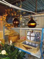 Photo showing wicker picnic baskets, multiple decorative glass vases with artificial flowers, and a flower wreath on a shelf, along with artificial plants below.