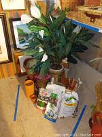 Faux peace lily plant in ceramic pot surrounded by bags of soil, stone chips, gardening tools, and terracotta pots.