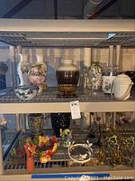 Shelf with various vases, glass bottles, decorative ceramic bowl, and small figurines including a bird; also metal wall sconces and artificial flowers on lower shelf.