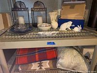 Full lot view showing candle holders, figurines, vases, plates, blankets, and linens on metal shelving rack.