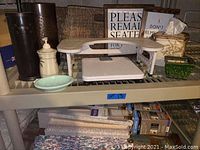 Shelf with wicker basket, tissue holder, toilet seat riser, dark metal cylindrical holders, ceramic soap dispenser, and other items arranged.