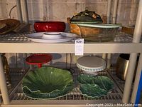 Various decorative serving dishes, bowls, platters, and ceramic items displayed on shelves including green leaf platters, a glazed basket with lid, and round cake holders.