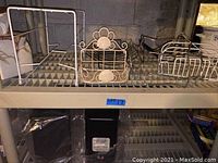Three metal wire baskets arranged on a wire shelf. One basket is white with a flower design, one is white rectangular, and one is a silver rectangular basket.