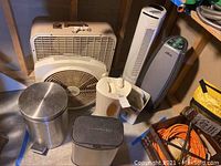 Photo showing various fans (box fan, tower fans) along with metal trashcans and a small humidifier.