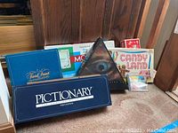 Photo showing boxed vintage board games including Pictionary First Edition game in blue box, Trivial Pursuit blue game box, Monopoly box with classic design, Candy Land game box with colorful logo, Scrabble Crossword Cube Game, UBI, and a triangular box with an eye graphic representing the Pictionary computer game.