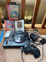 Sega Genesis console with controller, power adapter, and three game boxes: Sonic 3, Sonic Spinball, FIFA International Soccer arranged on a carpeted floor.