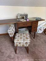 Right side angle of wooden sewing cabinet with fold-out side extensions, Kenmore model 1410 sewing machine visible, sewing accessories atop cabinet, paired with vintage floral upholstered sewing chair